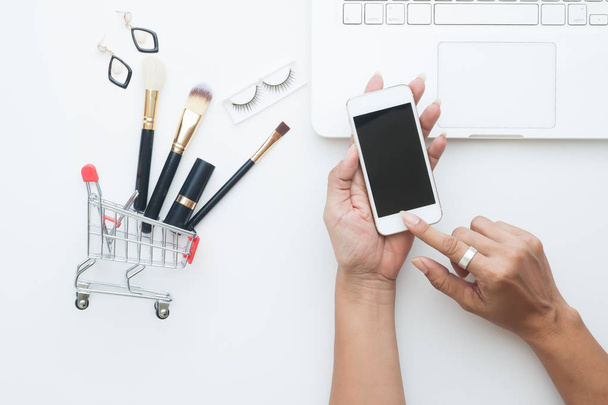 Елементи краси, пензлі, встановлені в кошик для покупок з рукою жінки, що тримає мобільний телефон, вигляд зверху концепція покупок онлайн
 - Фото, зображення