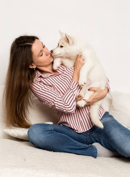 Happy beautiful woman hugging puppy husky. girl sitting on a sofa and puppy licks her - Foto, afbeelding