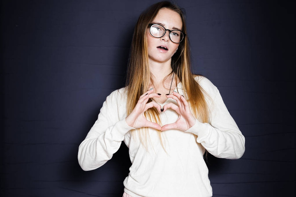 smiling girl in white sweater makes heart from fingers on a black background - Фото, изображение