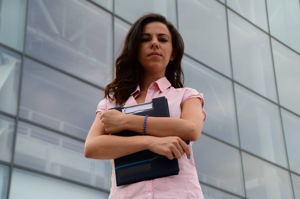 Νέοι όμορφη ευρωπαϊκών επιχειρήσεων γυναίκα στέκεται μπροστά από ένα γυάλινο κτίριο, κρατώντας σημειώσεις στα χέρια της, ψάχνει δεξιά στη φωτογραφική μηχανή - Φωτογραφία, εικόνα
