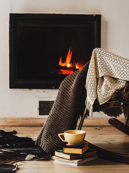 Silla mecedora con alfombra de punto, libros y taza de té o café antes de la chimenea
 - Foto, Imagen