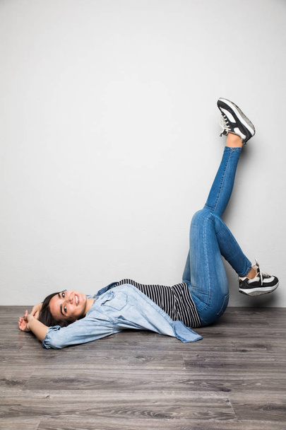 Šťastná neformální žena ležící na podlaze s nohama na vyvýšené nad šedým pozadím a při pohledu na fotoaparát - Fotografie, Obrázek
