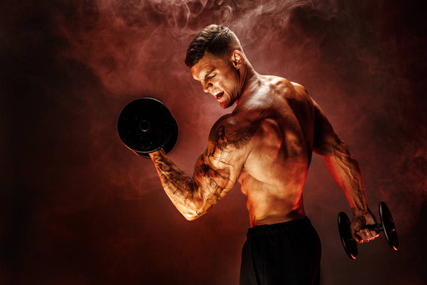 Strong sportsman lifting heavy dumbbell in smoke - Фото, изображение