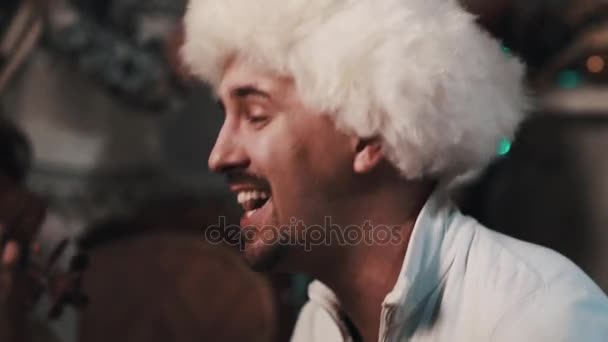 Αξύριστο αοιδός σε άσπρο fur καπέλο τραγουδώντας με κουαρτέτο εγχόρδων, κάθεται στο δωμάτιο - Πλάνα, βίντεο