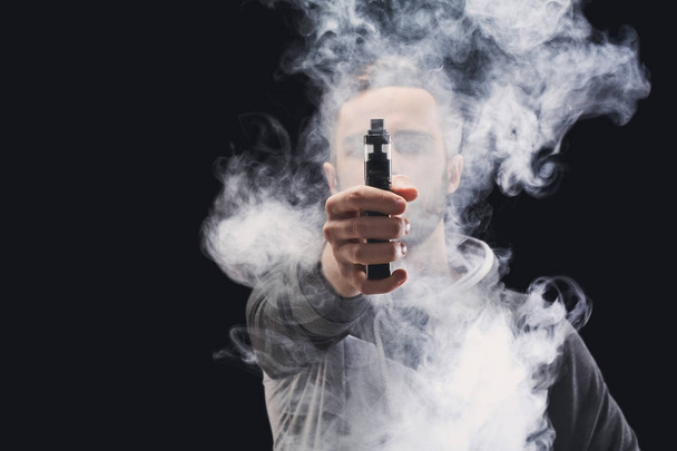 Young man vaping e-cigarette with smoke on black - Фото, зображення