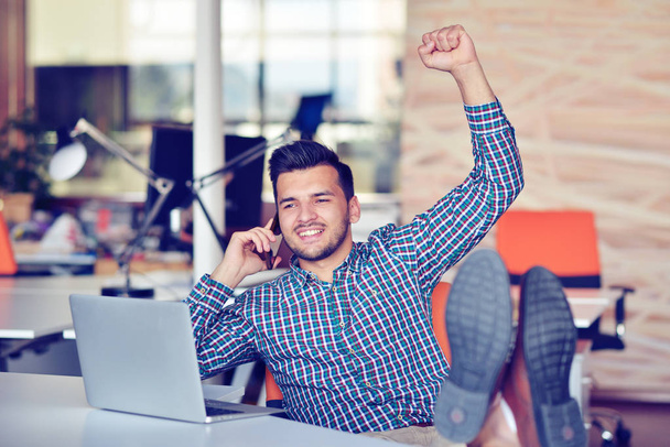 Cheerful young man in casual wear keeping arms raised and looking happy while sitting at the desk in office - Foto, Bild