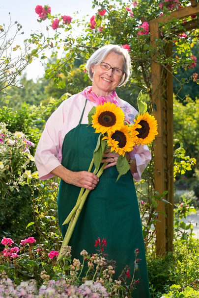 Hymyilevä vanhempi nainen pitelee auringonkukkia puutarhassa aurinkoisena päivänä
 - Valokuva, kuva