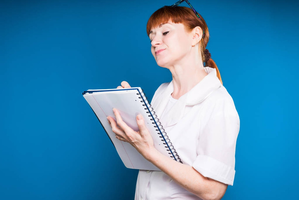 red-haired girl in medical clothes writes in a notebook of a record, isolated on a blue background - Photo, Image
