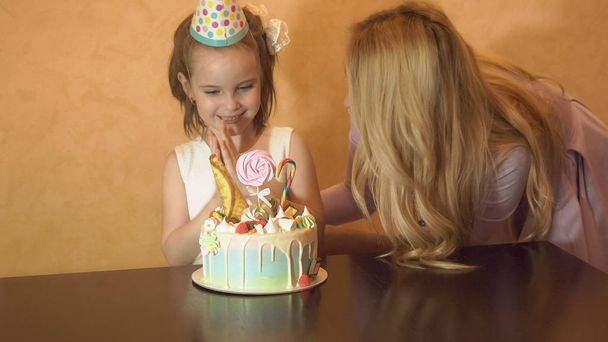 matka a dcera sfouknout svíčky na narozeninový dort. narozeniny malé holčičky. Oslavenkyně v slavnostní klobouk na dětské párty - Fotografie, Obrázek