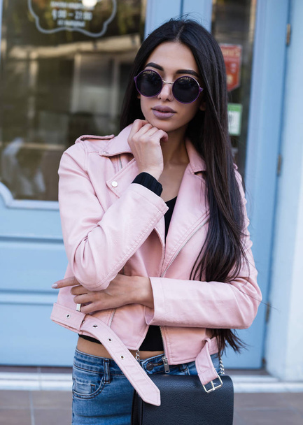 Street fashion concept - portrait of a pretty girl,walking in the street outside, Wearing pink leather jacket,grey sweater,big pink bag,sunglasses.Soft warm vintage color tone.Sunny weather. - Foto, afbeelding