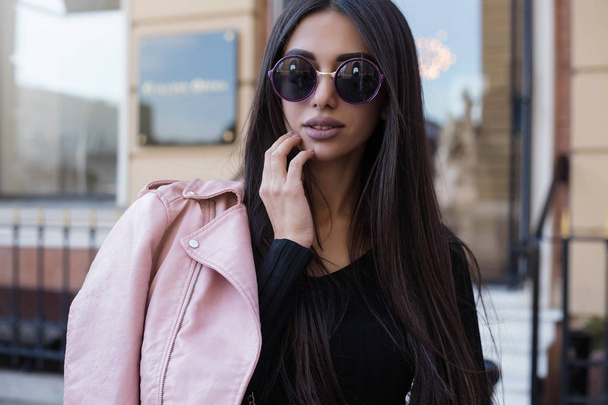 Fashion long hair woman with black shirt and hold pink leather jacket at city street. Stylish young model girl in elegant clothes outdoors. Lady with bright makeup.Fashion street style. - Photo, image