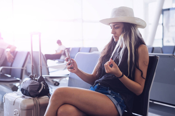 Flughafenfrau mit Hut auf Smartphone am Gate wartet im Terminal. Flugreise-Konzept. Etwas im Mobil suchen, App verwenden - Foto, Bild