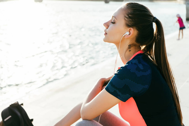 the girl rests after jogging and listens to music on headphones, on the river bank - Foto, imagen