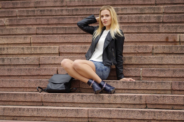 Beautiful blonde woman in a denim skirt and black leather jacket sits on the steps - Photo, Image