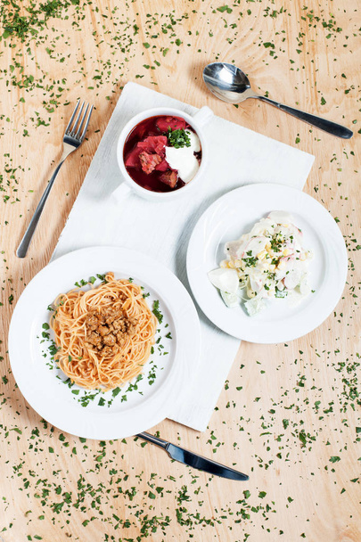 блюда из макарон, овощей, крабовых палочек, зелени и яиц на белых тарелках на белой разделочной доске на деревянном фоне в окружении зелени
 - Фото, изображение