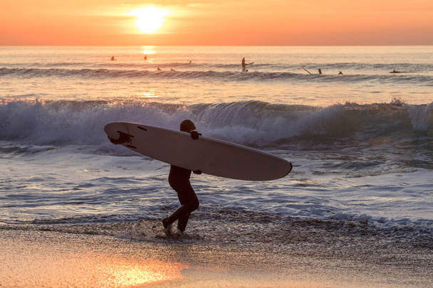 Surfer που μεταφέρουν την σανίδα του surf στο νερό με τα κύματα στο ηλιοβασίλεμα - Φωτογραφία, εικόνα
