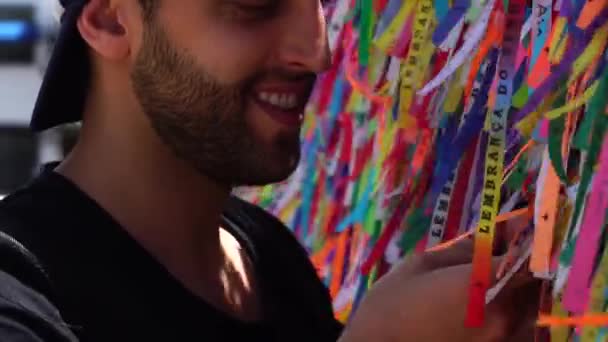 Guy making a wish with brazilian ribbons (Fita do Bonfim) on church fence in Salvador, Bahia, Brazil - Footage, Video
