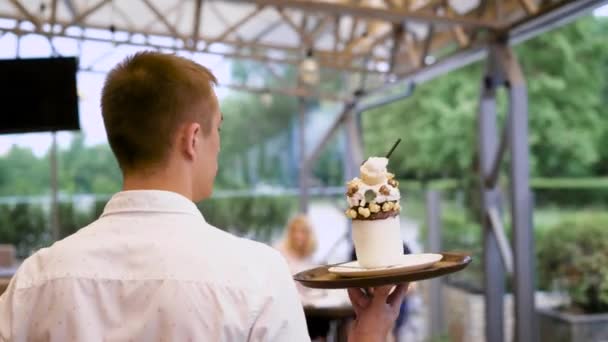 Hospitable waiter walking to the table bringing tasty dessert with whipped cream and chocolate for little client. Happy boy sitting in restaurant with parents and expressing joy having his pudding. - Filmati, video