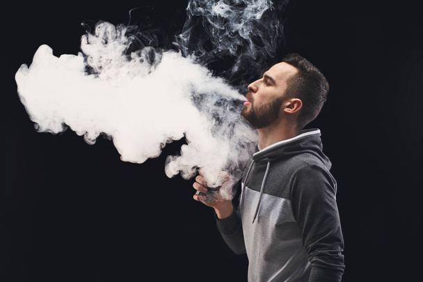 Young man vaping e-cigarette with smoke on black - Foto, afbeelding