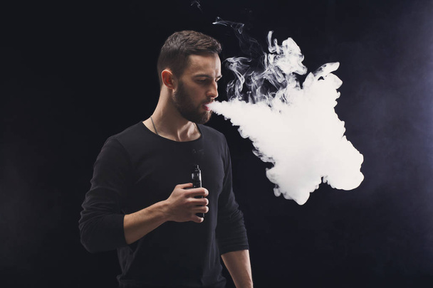 Jeune homme vapotage e-cigarette avec de la fumée sur noir
 - Photo, image