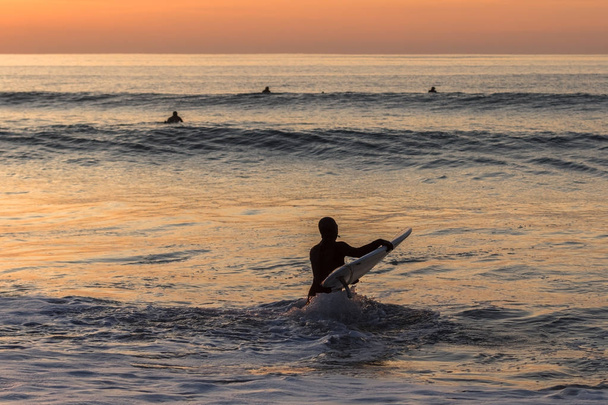 夕暮れの波と水の中にサーフボードを運ぶサーファー - 写真・画像