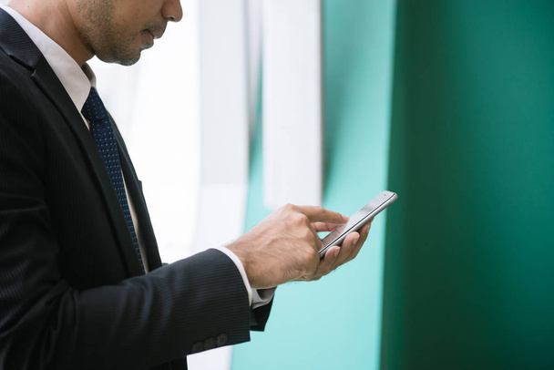 Бизнесмен, использующий приложение для мобильного телефона, пишет смс вне офиса в городе с небоскребами на заднем плане. Молодой азиат держит смартфон для деловой работы
. - Фото, изображение
