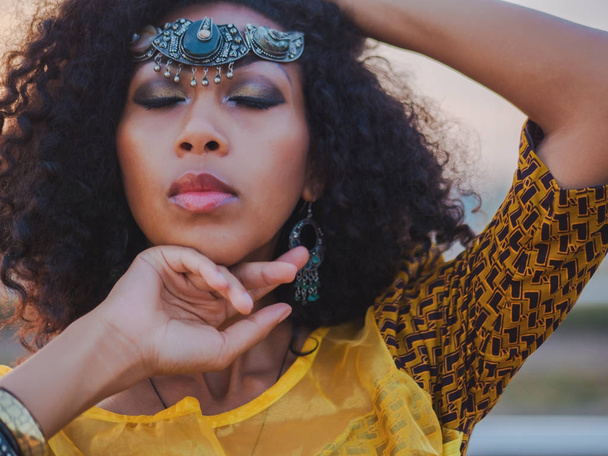 Portrait of young afro-american gypsy woman in colorful yellow traditional dress and silver crown on forehead smiling and dancing close to camera. Sexy fashion girl with curly hair - 写真・画像