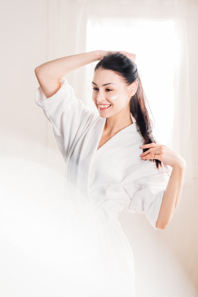 Lachende vrouw in badjas met crème op gezicht - Foto, afbeelding