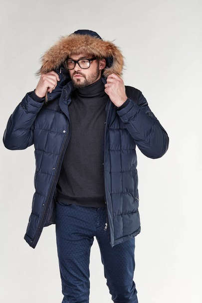 Attractive young sexy man model with beard and glasses posing in dark blue warm jacket with hood with a feather, and dark blue pants in light studio  - 写真・画像