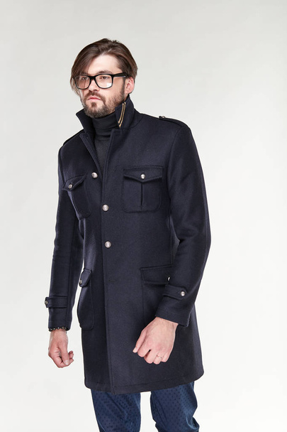 Attractive young sexy serious man model with beard and glasses posing in black cashmere coat and dark blue pants in light studio  - 写真・画像