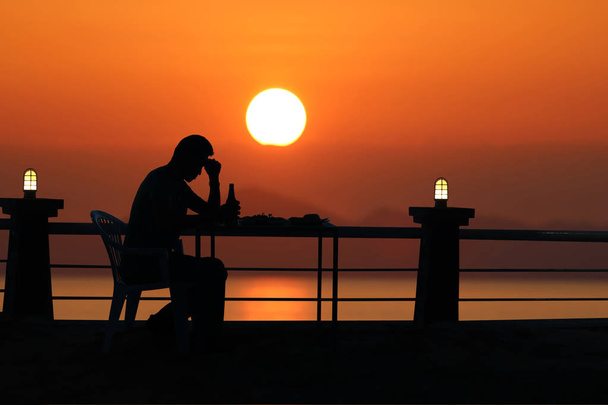 Силуэт грустный человек пьет пиво на пляже с красным небом солнца
 - Фото, изображение