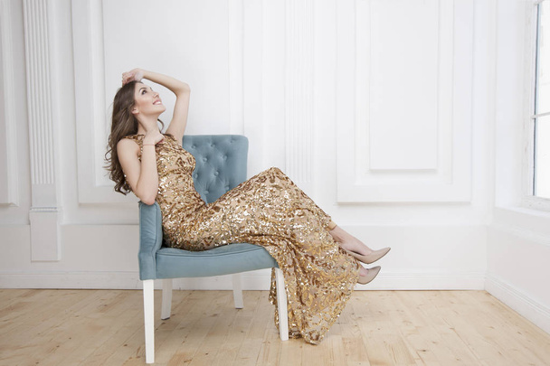 Young beauty woman in golden dress sitting in blue armchair, luxury apartments interior background  - Foto, afbeelding