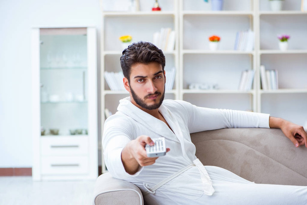 Young man in a bathrobe watching television at home on a sofa co - Photo, Image