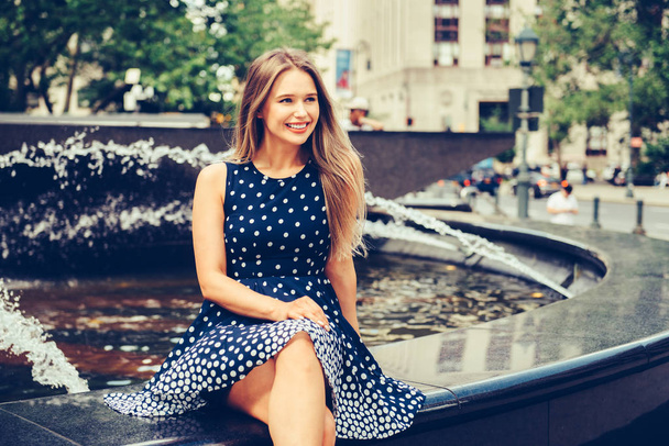 Beautiful young smiling woman sitting on city park fountain at summer day wearing dress and looking to the side  - Fotografie, Obrázek