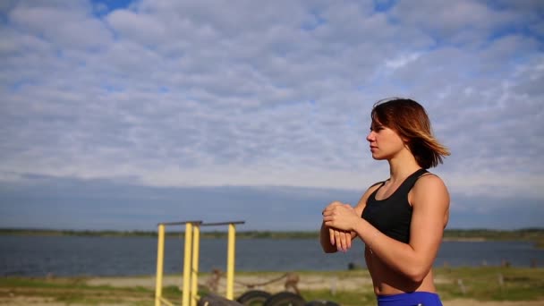 Sporty brunette girl sitting on a log by the river and conduct warm-up the hands before strength training - Footage, Video