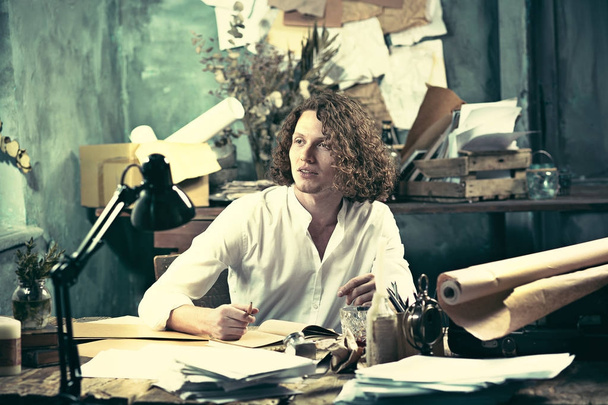 Writer at work. Handsome young writer sitting at the table and writing something in his sketchpad - Фото, изображение