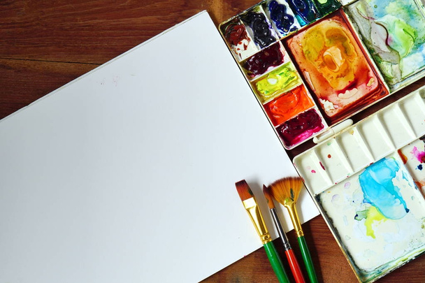 Palette von Aquarell mit Pinsel verschiedener Größe und weißem Papier auf hölzernem Texturhintergrund. - Foto, Bild