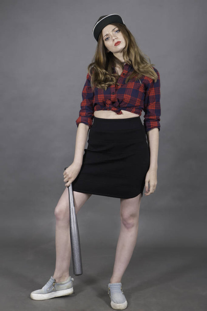 Young attractive woman dressed in baseball cap, red-blue plaid shirt and black skirt holding down pearl-grey bat in hand and looking to camera over the grey background - Zdjęcie, obraz