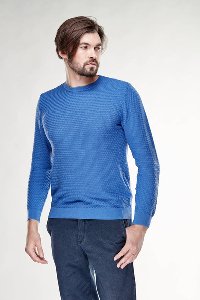 Attractive young sexy man model with beard and dark hair posing in blue sweater and dark blue pants in light studio  - 写真・画像
