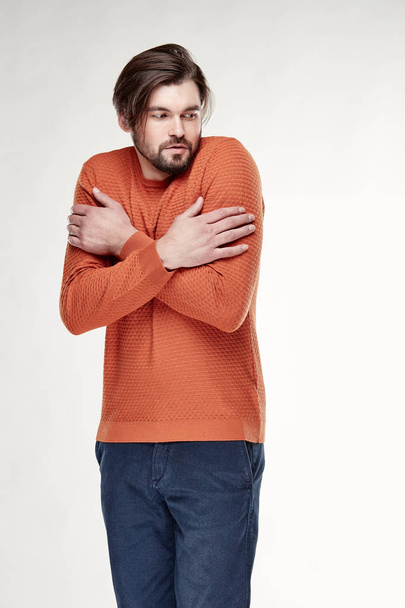 Attractive young sexy man model with beard and dark hair posing in orange sweater and dark blue pants in light studio  - 写真・画像