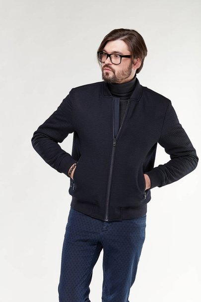 Attractive young sexy man model with beard and glasses posing in dark jacket with zipper and dark blue pants in light studio  - Photo, Image