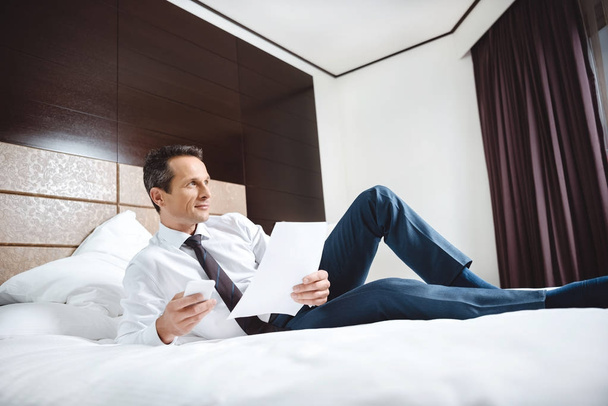 hombre de negocios en la cama con papeleo y teléfono inteligente
 - Foto, Imagen