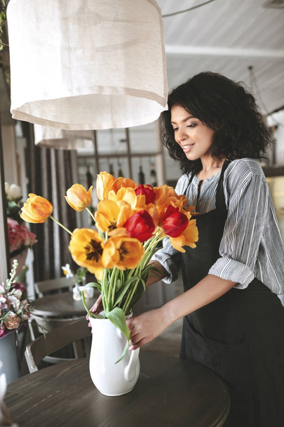 Young African American girl standing near flowers on table. Pretty florist in apron creating bouquet of tulips - Foto, Imagem