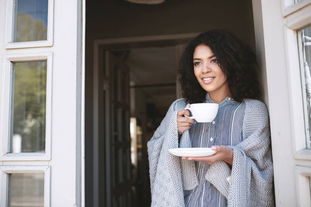 junges afrikanisch-amerikanisches Mädchen mit dunklem lockigem Haar, das an der Tür lehnend Kaffee trinkt. schöne Dame, die mit Tasse in der Hand in Plaid gewickelt steht - Foto, Bild