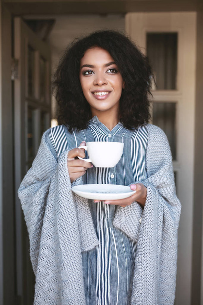 Молодая афроамериканка, стоящая с чашкой кофе, завернутой в клетку. Красивая девушка с темными вьющимися волосами пьет кофе
 - Фото, изображение