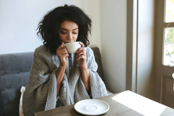 Beautiful African American girl drinking coffee in restaurant. Portrait of young lady with dark curly hair dreamily closing her eyes with cup in hands - 写真・画像