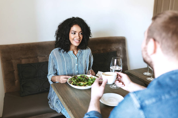 Όμορφη κοπέλα χαμογελαστή κάθεται στο εστιατόριο με τον φίλο και τρώγοντας σαλάτα. Αρκετά αφρικανική αμερικανική κυρία τρώγοντας σαλάτα στο γεύμα - Φωτογραφία, εικόνα