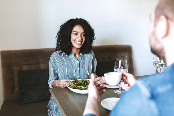 Красивая девушка сидит в ресторане с другом и ест салат. Улыбающаяся афроамериканка ест салат в кафе
 - Фото, изображение