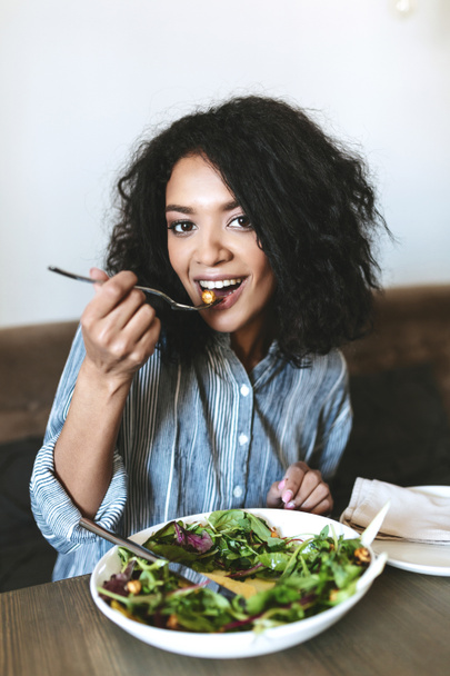 Nice African American girl eating salad in restaurant. Portrait of smiling lady with dark curly hair and salad on table at cafe - Photo, Image