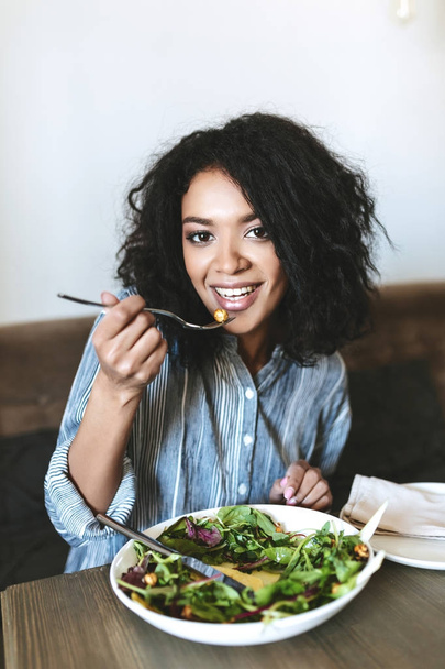 Pretty African American girl eating salad in restaurant. Portrait of smiling lady with dark curly hair and salad on table at cafe - Photo, Image
